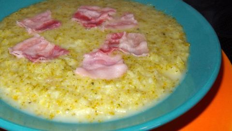 Eine cremige Brokkoli-Creme-Suppe in einer blauen Schüssel, garniert mit Schinkenstücken, auf einem orangefarbenen Untersetzer.