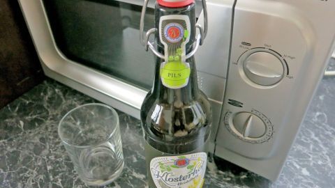 Warmes Bier gegen Sodbrennen: Erhitze vorsichtig eine Flasche in der Mikrowelle neben dem Glas. Dieser alte Hausmittel-Tipp könnte eine unerwartete Lösung für dein Problem sein. Bierflasche steht neben leerem Glas auf einer Marmoroberfläche, direkt vor einem TechnoStar-Mikrowellenofen.