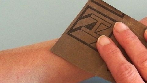 Eine Hand hält Schleifpapier und rubbelt damit über den Arm, um die Haarentfernung kostengünstig durchzuführen.
