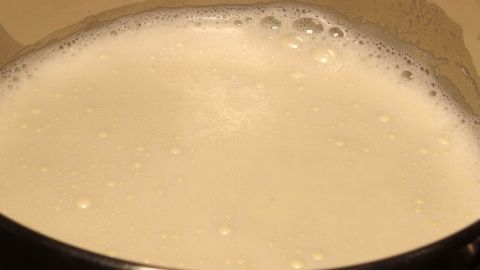 Übergekochte Milch auf dem Herd mit kalter Milch leicht entfernen. Ein Topf mit schäumender Milch auf einem Herd wird vom überschüssigen Schaum gereinigt. Das Küchenumfeld ist sichtbar.