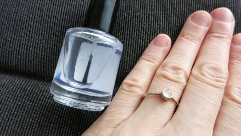 Ein Silberring liegt neben einem Nagellackfläschchen auf einem schlichten Untergrund, während eine Hand mit dem Ring die Fläche berührt.