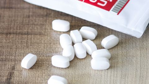 Lysin kann zur Bekämpfung und Vorbeugung von Herpes als Nahrungsergänzungsmittel eingenommen werden. Weiße Lysin-Tabletten liegen auf einem Holztisch, während die geöffnete Verpackung im Hintergrund sichtbar ist.