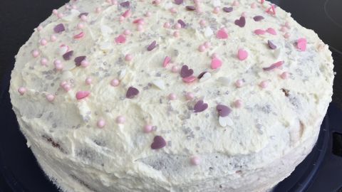 Eine Quark-Sahne-Obst-Torte mit einer cremigen, weißen Oberfläche und bunten Dekorationsstückchen steht auf einem blauen Teller.