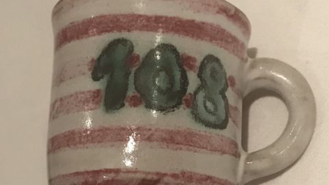 Ein kleiner Becher mit rot-weiß gestreiftem Muster und der Aufschrift "108", auf einem neutralen Untergrund platziert.