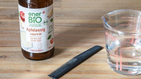 Eine Flasche mit Apfelessig und einem Kamm liegen neben einem Glas Wasser auf einem Holztisch, bereit zur Anwendung gegen Kopfläuse.