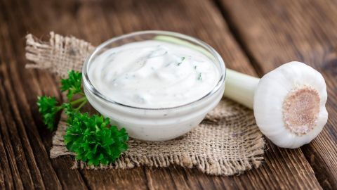 Eine Schüssel mit Knoblauch-Remoulade steht auf einem rustikalen Holzbrett, daneben liegt eine frische Knoblauchknolle und Petersilie.