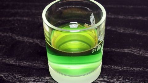 Ein Glas mit mehrfarbigem Getränk steht auf einer dunklen Holzoberfläche, wobei grüne und klare Schichten sichtbar sind.