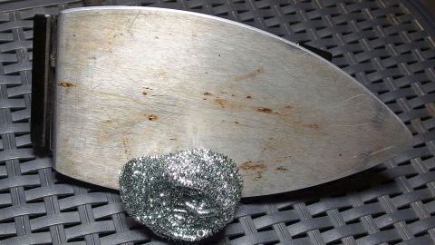 Ein schmutziges Bügeleisen mit einer mit Stahlwolle belegten Fläche liegt auf einem strukturierten, schwarzen Untergrund.
