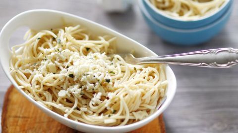 Eine Schüssel Spaghettisalat mit Kräutern und Käse steht auf einem Holzteller, während im Hintergrund eine kleine Schüssel mit Pasta zu sehen ist.