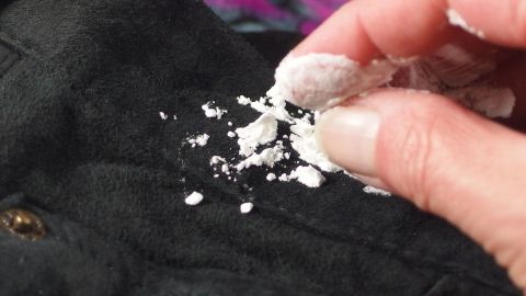 Wildlederjacke, besonders den Kragen, mit Speisestärke reinigen. Eine Hand streut Speisestärke auf einen schwarzen Wildlederkragen, um Flecken zu reinigen. Der Hintergrund ist verschwommen.