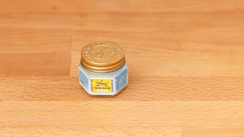 Eine kleine, sechseckige Dose mit goldener Verschlusskappe steht auf einem Holzuntergrund. Sie enthält die Chinasalbe gegen Kopfschmerzen.