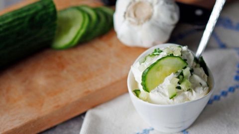 Eine Schüssel mit cremigem Tzatziki, garniert mit Gurkenscheiben, steht auf einem Tisch neben einer geschnittenen Gurke und Knoblauch.