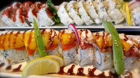 Sushirollen liegen auf einem Teller, garniert mit Avocado, Mango und Sesam, besprenkelt mit Soße, daneben Zitronenscheiben.