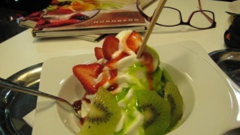 Joghurt mit frischen Erdbeeren und Kiwi, garniert mit rotem und grünem Sirup, auf einem Tisch neben einer Brille und Magazinen.