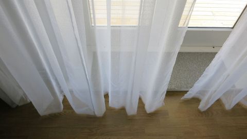 Durchsichtige Gardinen hängen vor einem Fenster und stehen im Fokus, während die Umgebung aus Holzfußboden besteht.