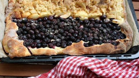 Ein Blech Selterskuchen mit einem Belag aus Äpfeln und Blaubeeren liegt auf einem Tisch, umgeben von einem karierten Tuch.