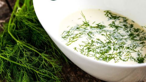 Eine weiße Schüssel enthält eine cremige Honig-Senf-Sauce, garniert mit frischem Dill, vor einer rustikalen Holzoberfläche.
