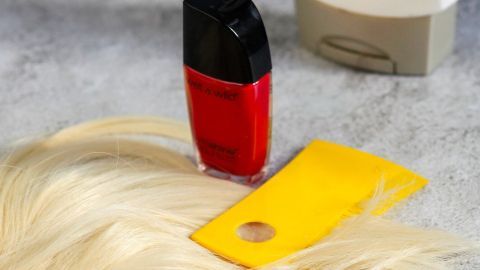 Ein roter Nagellack steht auf einem Tisch neben blonden Haaren und einem gelben Kamm in einem neutralen Raum.
