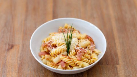 Ein Teller Penne mit Spargel-Carbonara steht auf einem Holztisch, überzogen mit Käse und frischen Kräutern.