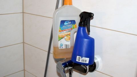 Eine blaue Sprühflasche und eine transparente Flasche hängen an einer Duschwand, bereit zur Reinigung der Badewanne und Duschtasse.
