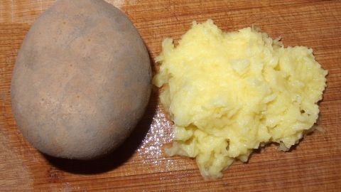 So erspart man sich das mühsame Reiben der rohen Kartoffeln: Kartoffeln schälen, kleinschneiden, in den Mixer geben, mit reichlich Wasser auffüllen, mixen und die Masse dann durch ein Leinentuch abseihen und auspressen. Eine rohe Kartoffel liegt neben geriebenen Kartoffeln auf einem Holzbrett, bereit für die Zubereitung von Klößen.