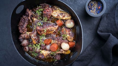 Im schwarzen Topf köchelt Coq au vin mit Hähnchen, Karotten und Zwiebeln, umgeben von frischen Kräutern und Gewürzen.