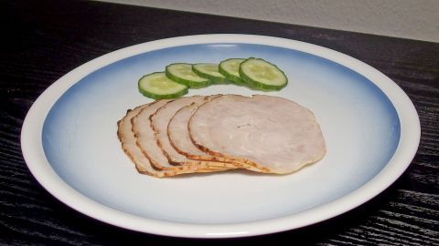 Scheiben von Bratenaufschnitt liegen ordentlich auf einem weißen Teller, umgeben von frischen Gurkenscheiben auf einem dunklen Tisch.