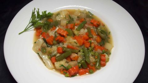 Eine Gemüsesuppe ist in einer weißen Schale angerichtet, mit Karotten, Bohnen und Kräutern, die appetitlich präsentiert ist.