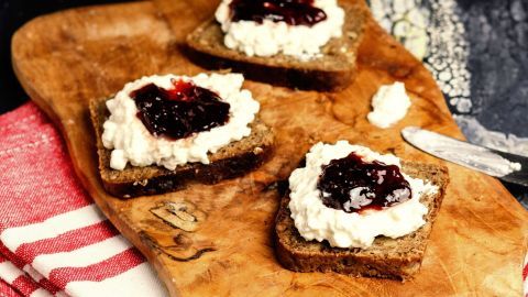 Hüttenkäse mit etwas Sauerrahm schmeckt super unter Marmelade auf dem Frühstücksbrot und ist ein guter Ersatz für Butter. Drei Brotscheiben mit feinem Aufstrich und Marmelade liegen auf einem Holzbrett, umgeben von Küchenutensilien.