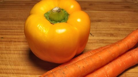 Eine gelbe Paprika liegt neben zwei Karotten auf einem Holzbrett, bereit zum schonenden Garen in der Küche.