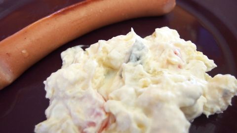 Wenn selbst gemachte Mayonnaise ein bisschen zu dickflüssig geworden ist, einfach mit etwas Wasser verflüssigen. Ein Klumpen Kartoffelsalat liegt auf einem Teller, umgeben von einer Wurst, die als Beilage dient, möglicherweise für ein BBQ.