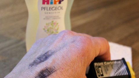 Einfach mit Babyöl und Klopapier schrubben und schon ist die Haut wieder sauber. Eine Hand mit Druckertinte zeigt Schmierflecken, während im Hintergrund eine Flasche Babyöl steht.