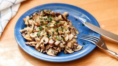 Auf einem blauen Teller liegt eine Champignonpfanne, die appetitlich zubereitete Pilze und Gewürze zeigt, in einer gemütlichen Umgebung.