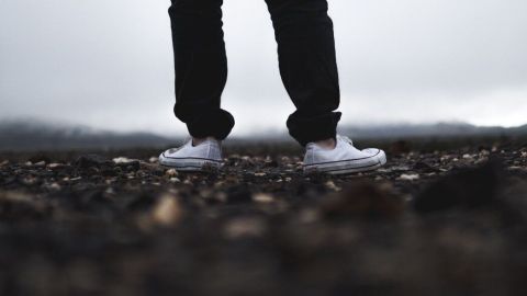 Weiße Stoffturnschuhe stehen auf einem schmutzigen Untergrund, während eine graue, wolkige Landschaft im Hintergrund sichtbar ist.
