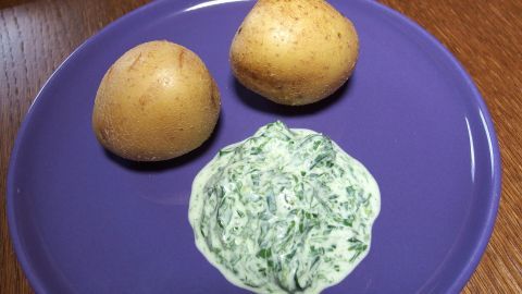 Zwei Pellkartoffeln liegen neben einem cremigen Dip auf einem lila Teller in einer holzgetäfelten Umgebung.