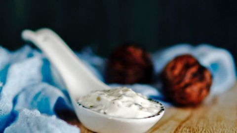 Ein cremiger Edel-Feta-Dip liegt in einem Löffel, umgeben von blauen Stoff und zwei dunklen, runden Snacks.