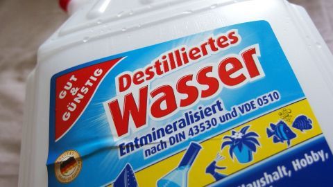 Eine Flasche mit destilliertem Wasser steht vor einem hellen Hintergrund und zeigt den Etikett mit Reinheitsgarantie und Anwendungshinweisen.