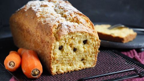 Saftiger Möhrenkuchen liegt auf einem Gitter, umgeben von frischen Karotten und einem Lila-Tuch im Hintergrund.
