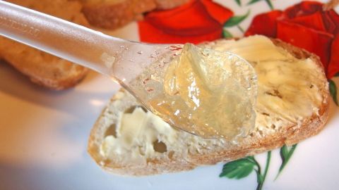 Ein Löffel Apfelgelee wird auf eine belegte Brotscheibe verteilt, umgeben von roten Rosenblättern auf einem Teller.