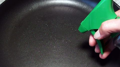 Eine Hand sprüht Speiseöl aus einer grünen Sprühflasche in eine schwarze Pfanne, die auf einer Arbeitsfläche steht.