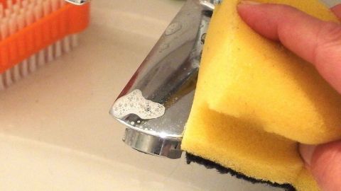 Eine Hand hält einen Schwamm und reinigt eine glänzende Armatur im Badezimmer, um sie fleckenfrei zu machen.