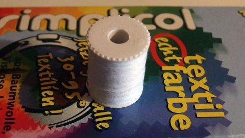 Weißes Garn beim Färben von Wäsche gleich mitfärben. Ein Spulen Garn liegt auf einer Packung Textilfarbe, bereit zum Färben für die Wäsche. Farbige Muster umgeben das Garn.