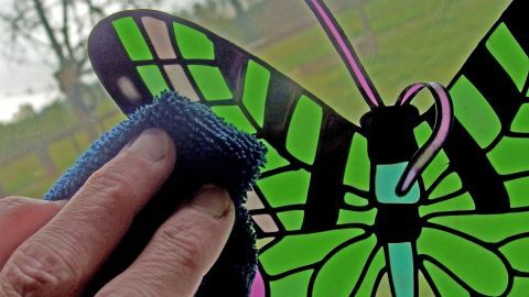 Mithilfe von Essig lassen sich alte Window-Color-Reste leichter lösen und abziehen. Ein Hand entfernt mit einem Tuch Fensterfarbe von einem bunten Schmetterlingsaufkleber, während im Hintergrund ein Feld sichtbar ist.