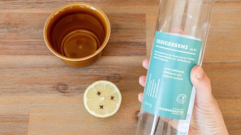 Eine Person hält eine Glasflasche über einer halben Zitrone mit Nelken. Im Hintergrund steht eine braune Schüssel auf einem Holzuntergrund.