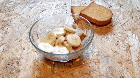 Eine Glasschüssel mit Zwieback-Brei und Bananenstücken steht auf einem Tisch, daneben zwei Stücke Zwieback.