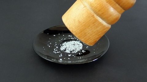 Eine Holzpfeffermühle streut Salz auf einen kleinen schwarzen Teller; kontrastreiche Darstellung auf dunklem Hintergrund.