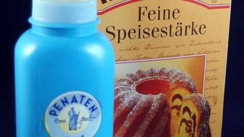 Eine blaue Flasche mit Babypuder steht neben einer Packung Speisestärke, die auf einem dunklen Untergrund platziert ist.