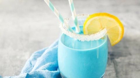 Ein blauer Cocktail mit zwei gestreiften Strohhalmen steht auf einem Tisch, umgeben von Eiswürfeln und einem blauen Tuch.