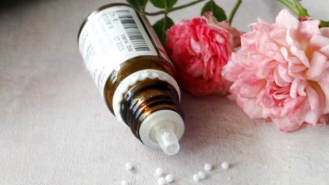 Ein Braunglasfläschchen mit Chinin, umgeben von rosa Blumen, liegt auf einer Tischoberfläche und hat einige Kügelchen daneben verstreut.