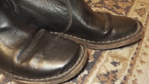 Schwarze Kunstlederstiefel stehen auf einem Musterteppich, zeigen Flecken durch Imprägnierspray auf der Oberfläche.